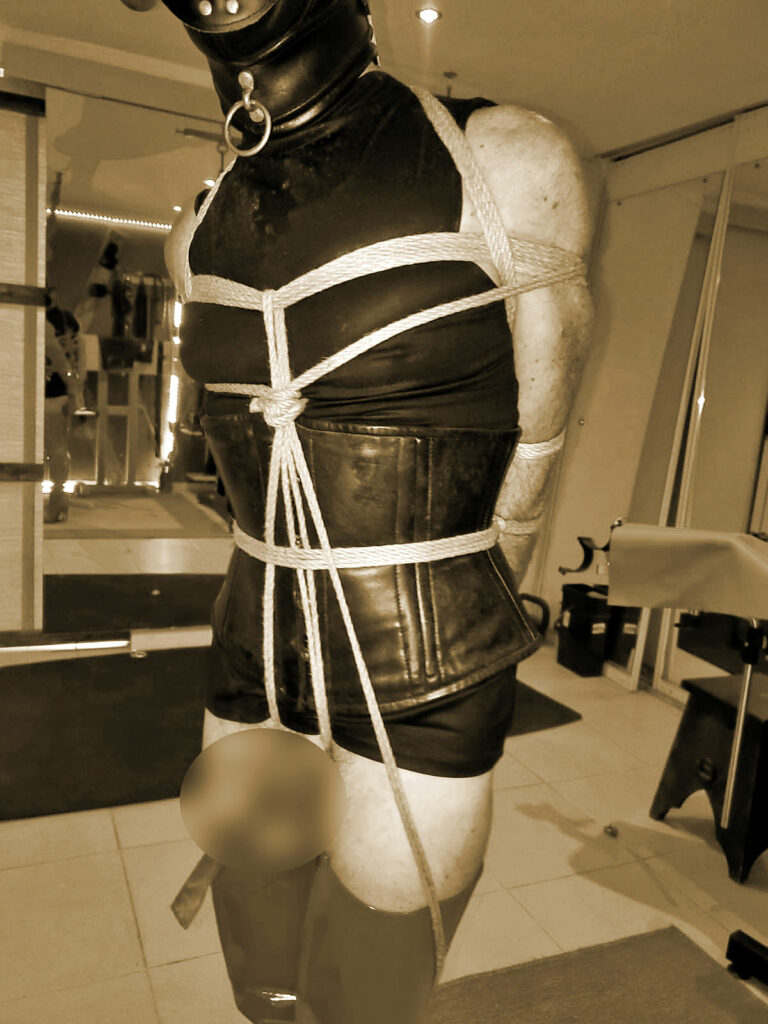 Esclave portant cagoule, corset et cuissardes attaché en position verticale